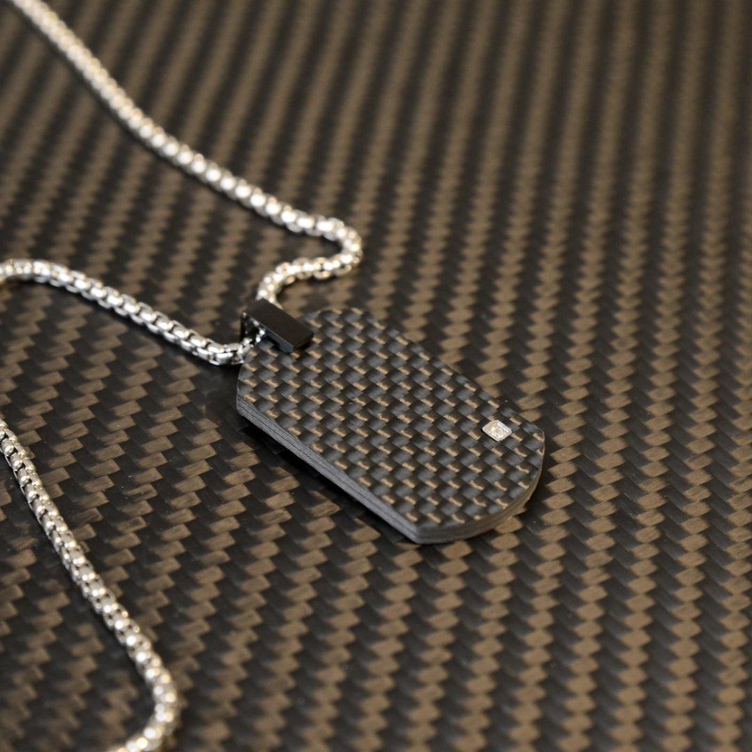 Close-up of a 3K carbon fiber oval pendant with a checkered pattern and a small diamond accent, paired with a silver-tone chain, displayed on a carbon fiber textured background.