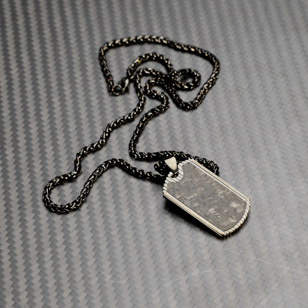 Close-up of a 3K carbon fiber dog tag pendant with metallic accents, paired with a black-tone chain, displayed on a carbon fiber textured background.