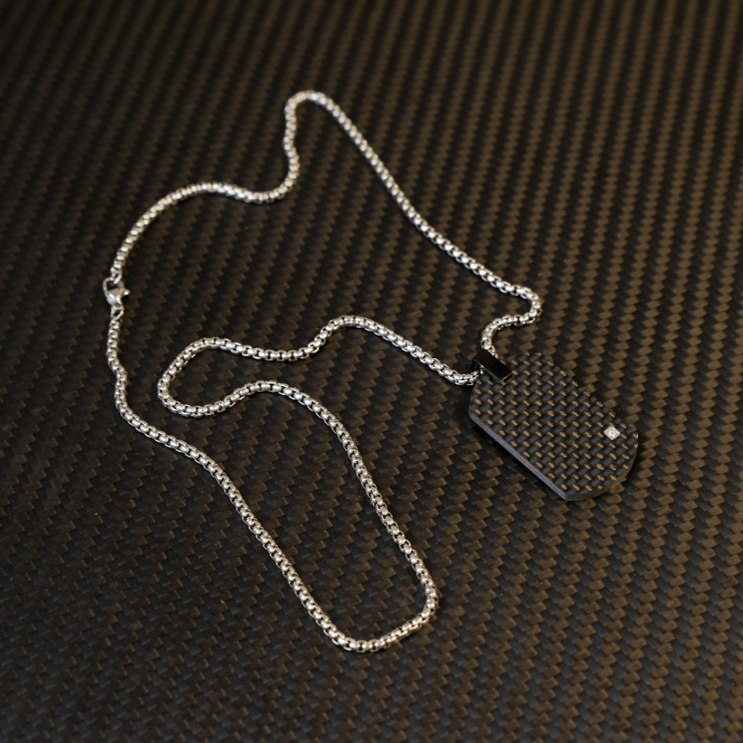 Close-up of a 3K carbon fiber dog tag pendant with a checkered pattern and a small diamond accent, paired with a silver-tone chain, displayed on a carbon fiber textured background.