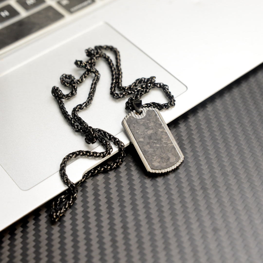 Close-up of a 3K carbon fiber dog tag pendant with metallic detailing, paired with a black-toned chain, displayed on a laptop and a carbon fiber textured background.