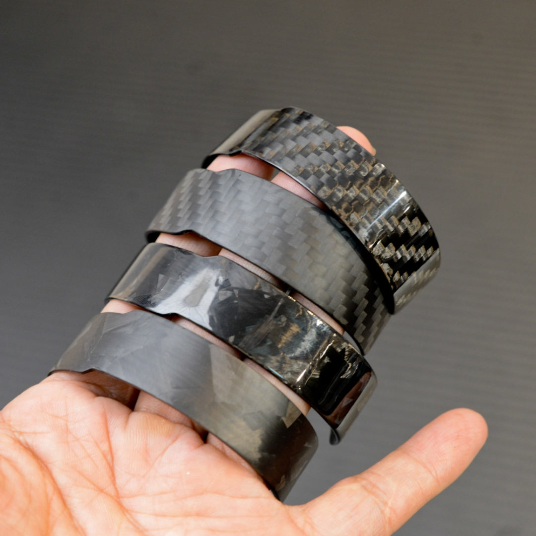 A hand holding four black carbon fiber bracelets with distinct weave patterns, displayed against a dark background.