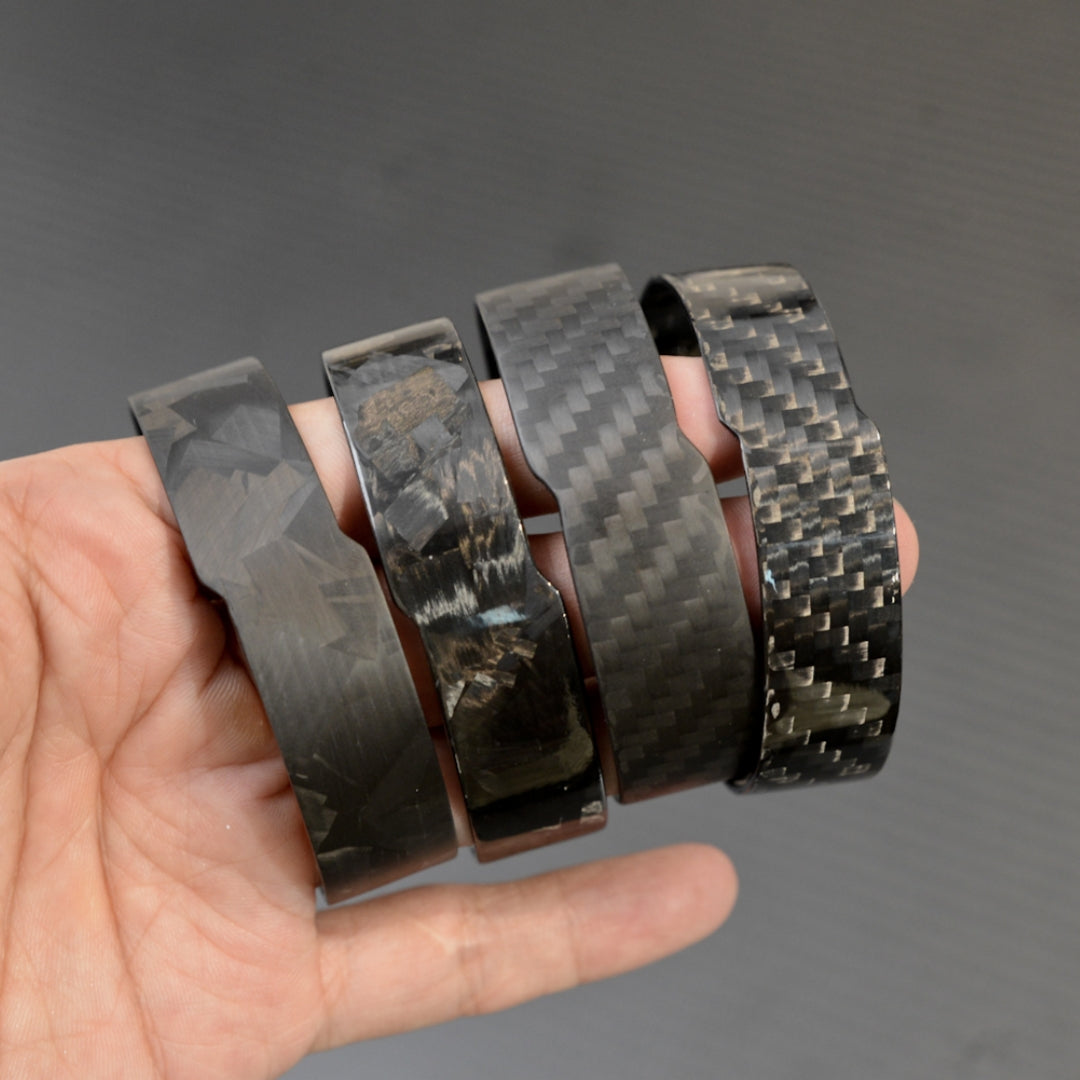 A hand holding four black carbon fiber bracelets with distinct weave patterns, displayed against a dark background.