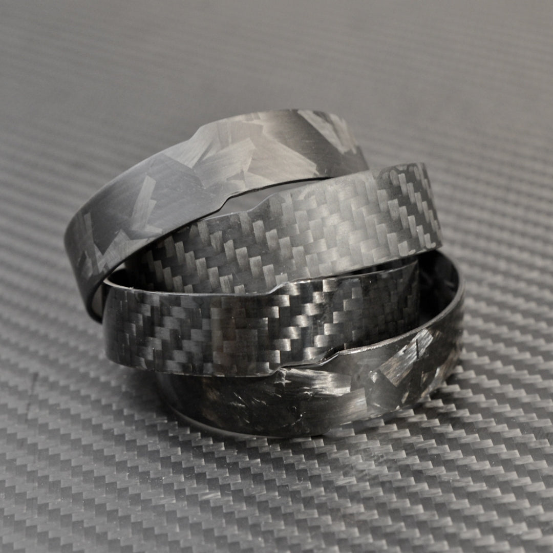 Four carbon fiber cuff bracelets with distinct weave patterns, stacked on a carbon fiber textured surface.