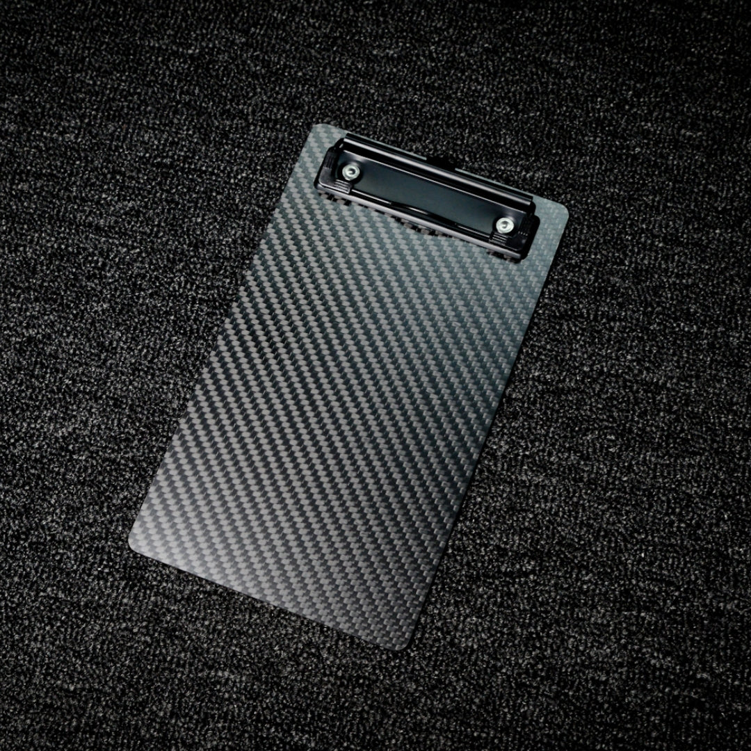Close-up detail of a 3K carbon fiber clipboard featuring a checkerboard pattern and a black metal clip, resting on a textured dark surface.