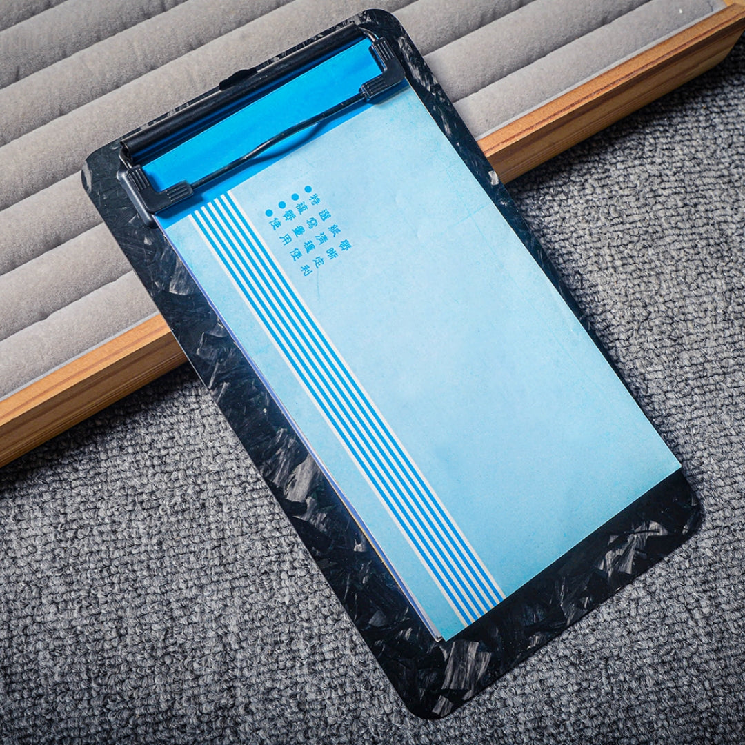 Close-up detail of a forged carbon fiber clipboard with a black metal clip, holding blue paper with Chinese characters, placed on a textured surface with a wooden and padded background.