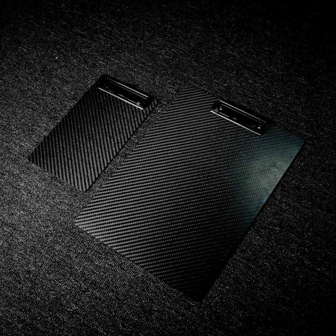 Close-up detail of two 3K carbon fiber clipboards with a distinct woven pattern and black metal clips, placed on a dark textured surface.