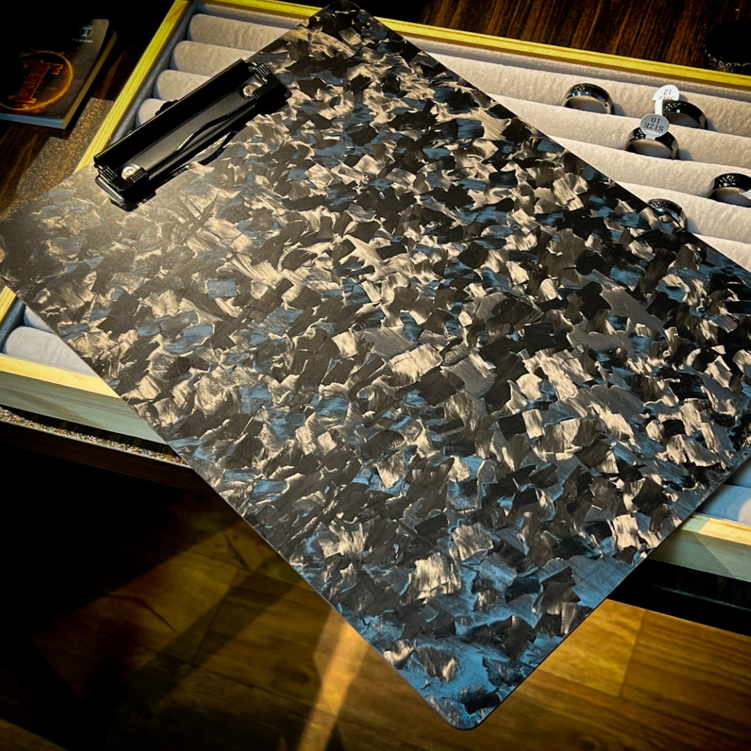 Close-up detail of a forged carbon fiber clipboard with a distinct chopped pattern and glossy metallic texture, equipped with a black metal clip, placed over a display case with jewelry.