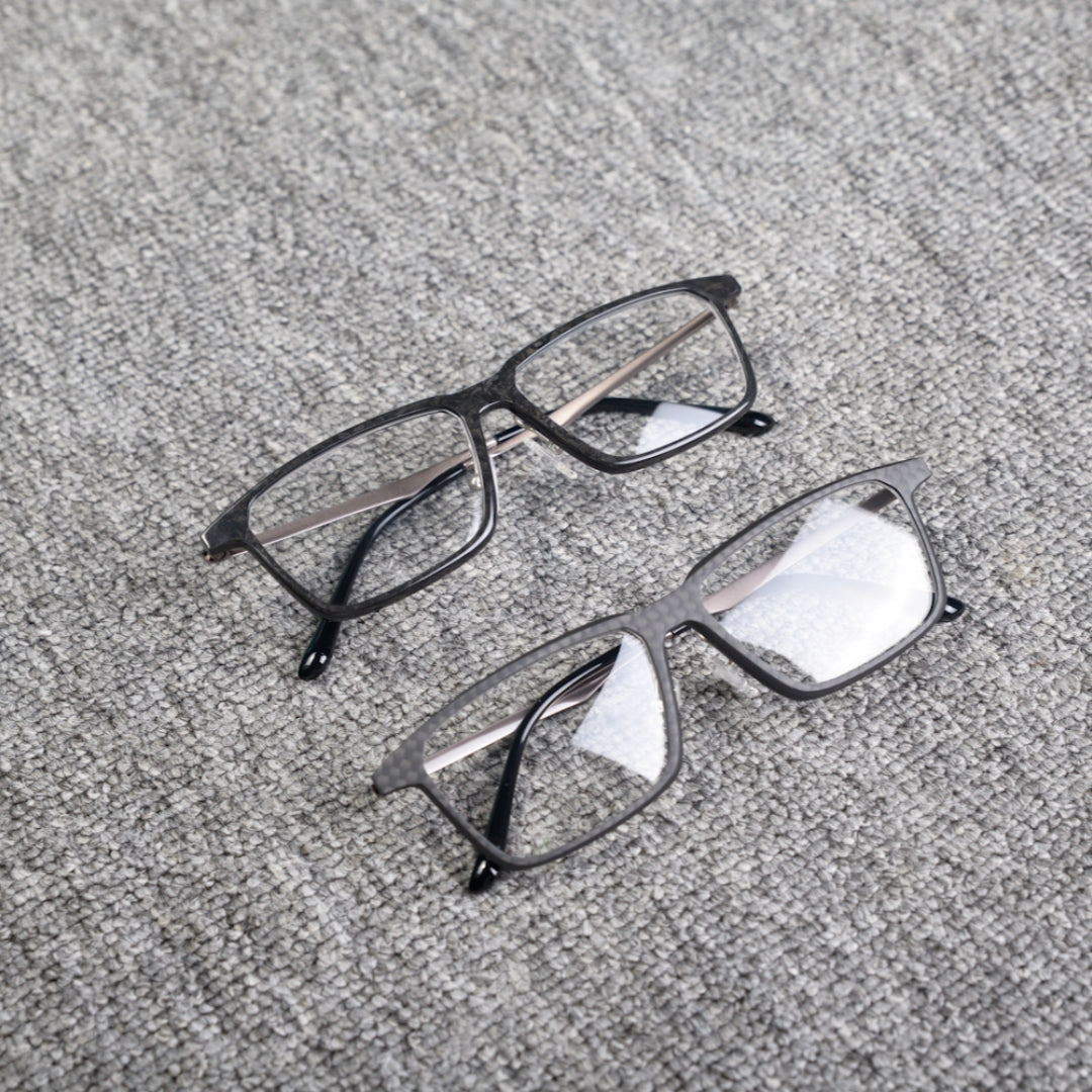 Close-up of two eyeglass frames made from forged carbon fiber and 3K carbon fiber, featuring sleek black designs with clear lenses, placed on a textured gray surface.