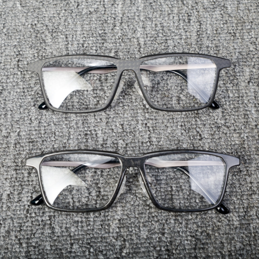 Close-up of two pairs of eyeglass frames crafted from forged carbon fiber and 3K carbon fiber, showcasing sleek black designs with clear lenses, placed on a textured gray surface.