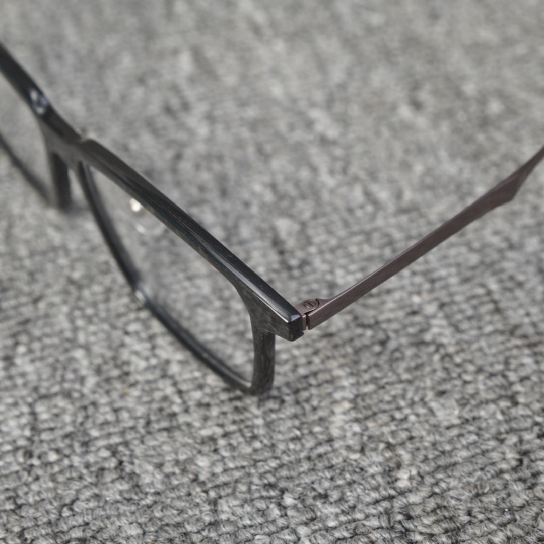 Close-up side detail of a forged carbon fiber eyeglass frame, showcasing the sleek black frame and metallic temple connection, placed on a textured gray surface.