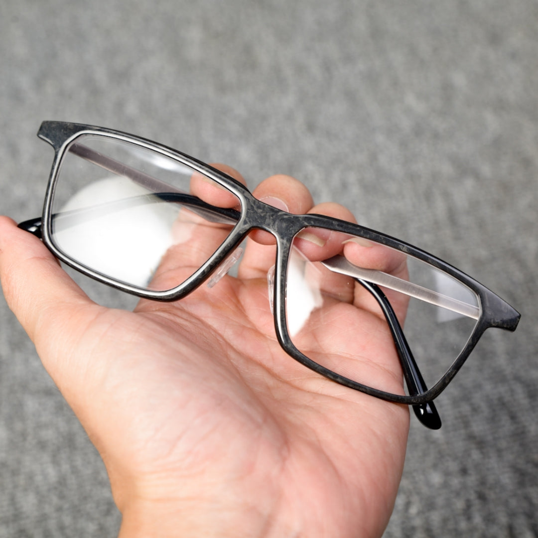 Close-up of a forged carbon fiber eyeglass frame with a sleek black design and clear lenses, held in a hand against a textured gray background.