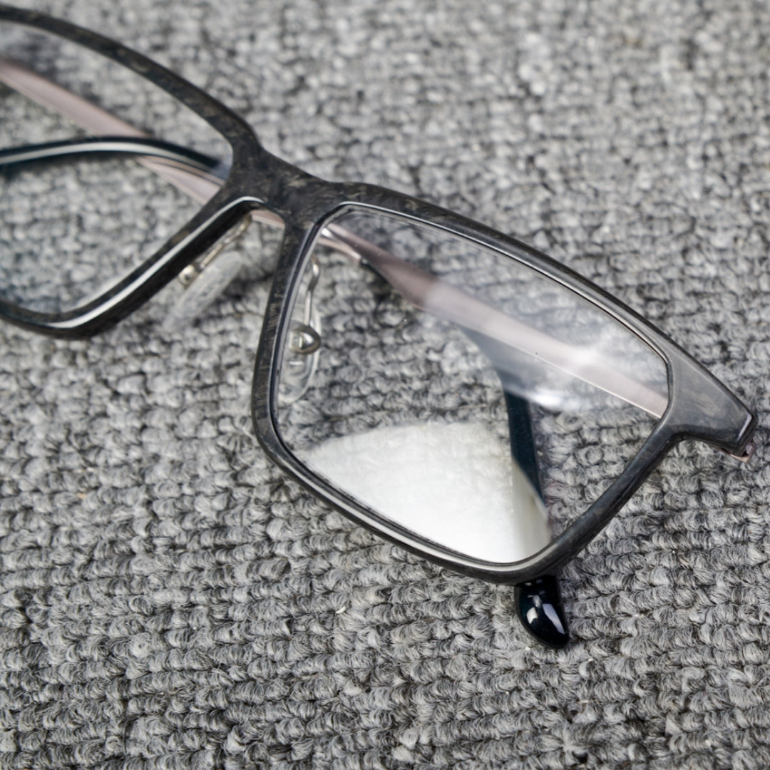 Close-up detail of a forged carbon fiber eyeglass frame with a sleek black design and clear lenses, placed on a textured gray surface.