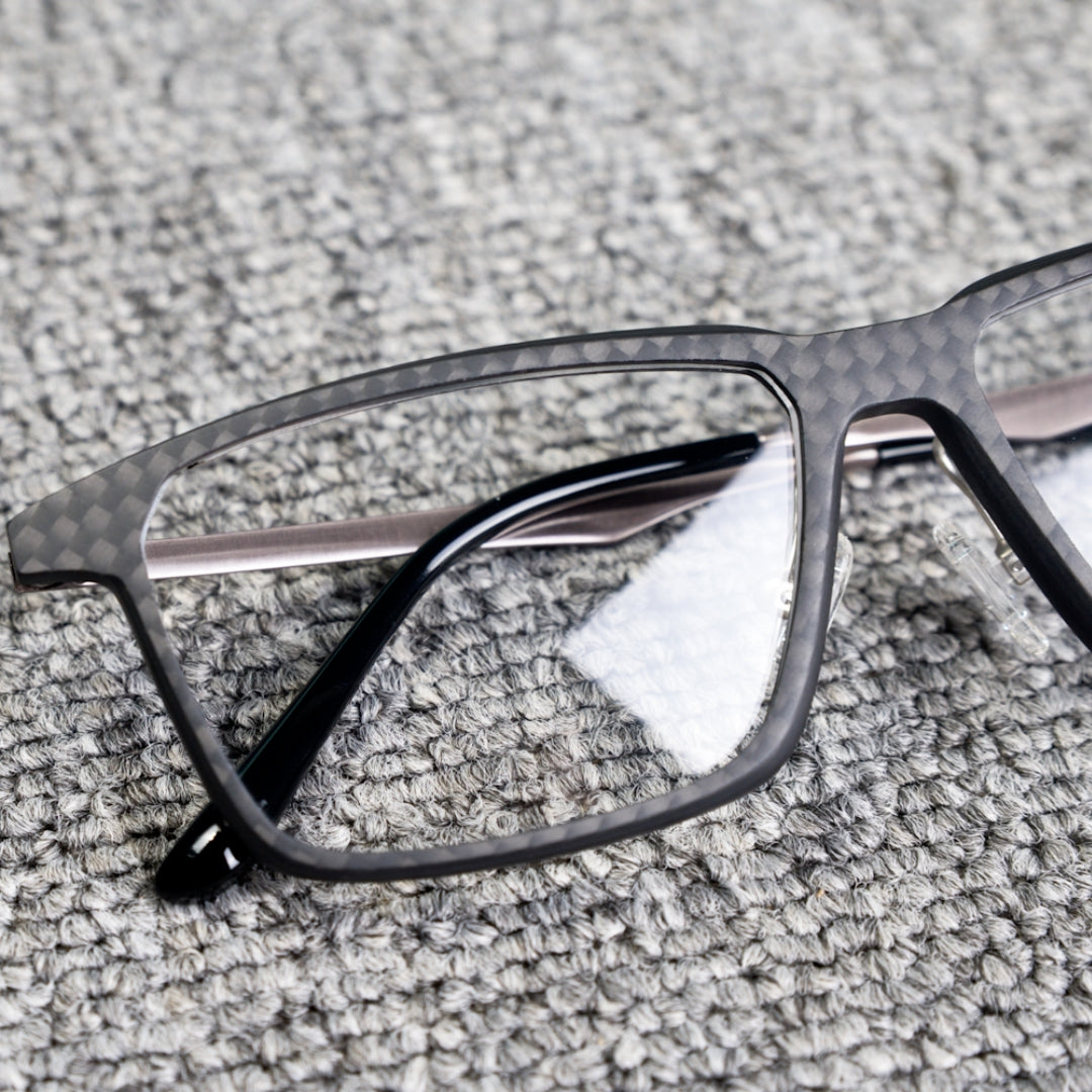 Close-up detail of a 3K carbon fiber eyeglass frame featuring a sleek black textured pattern, metallic temples, and clear lenses, placed on a textured gray surface.