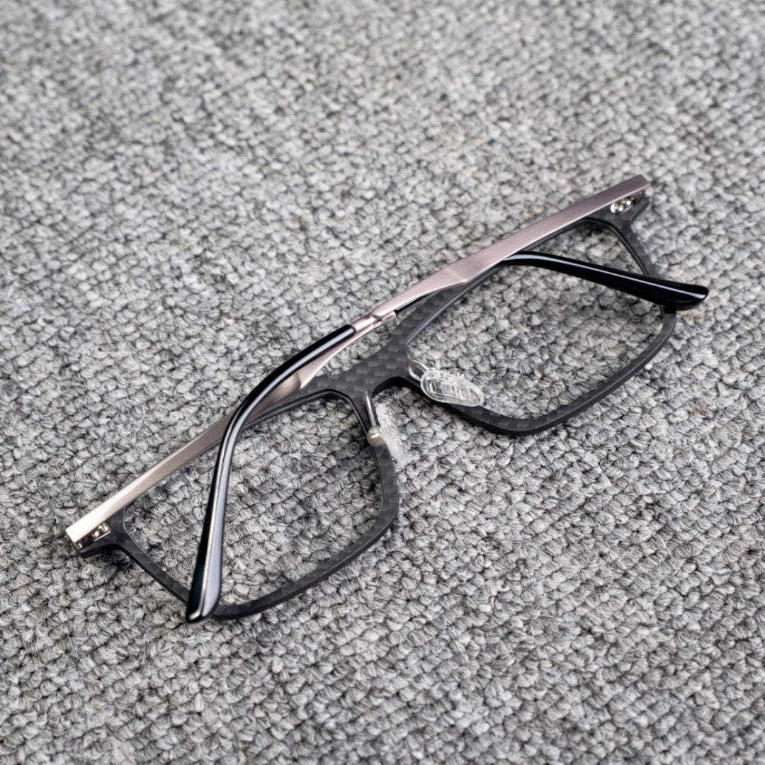 Close-up reverse side detail of a 3K carbon fiber eyeglass frame with a sleek black textured pattern and metallic temples, placed on a textured gray surface.