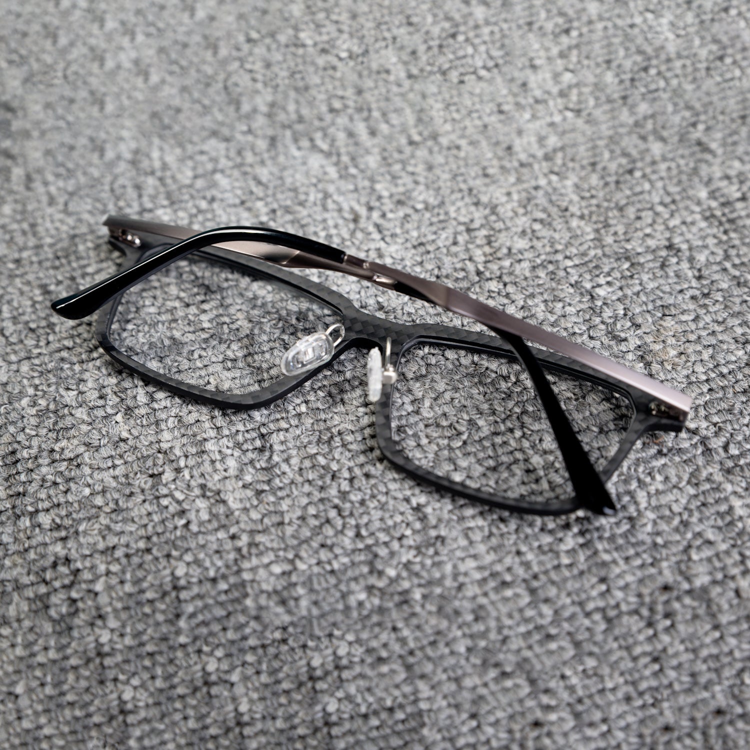 Close-up reverse side detail of a 3K carbon fiber eyeglass frame, showcasing its sleek black textured pattern and metallic temples, placed on a textured gray surface.
