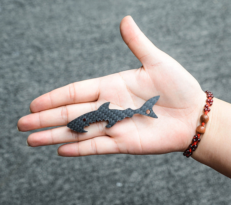 Close-up of a shark-shaped 3K carbon fiber keychain with a checkered pattern and serrated edge details, resting on an open palm adorned with a red and brown beaded bracelet, against a gray textured background.