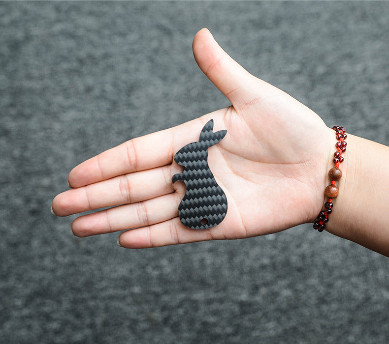 Close-up of a rabbit-shaped 3K carbon fiber keychain with a checkered pattern, resting on an open palm adorned with a red and brown beaded bracelet, against a gray textured background.