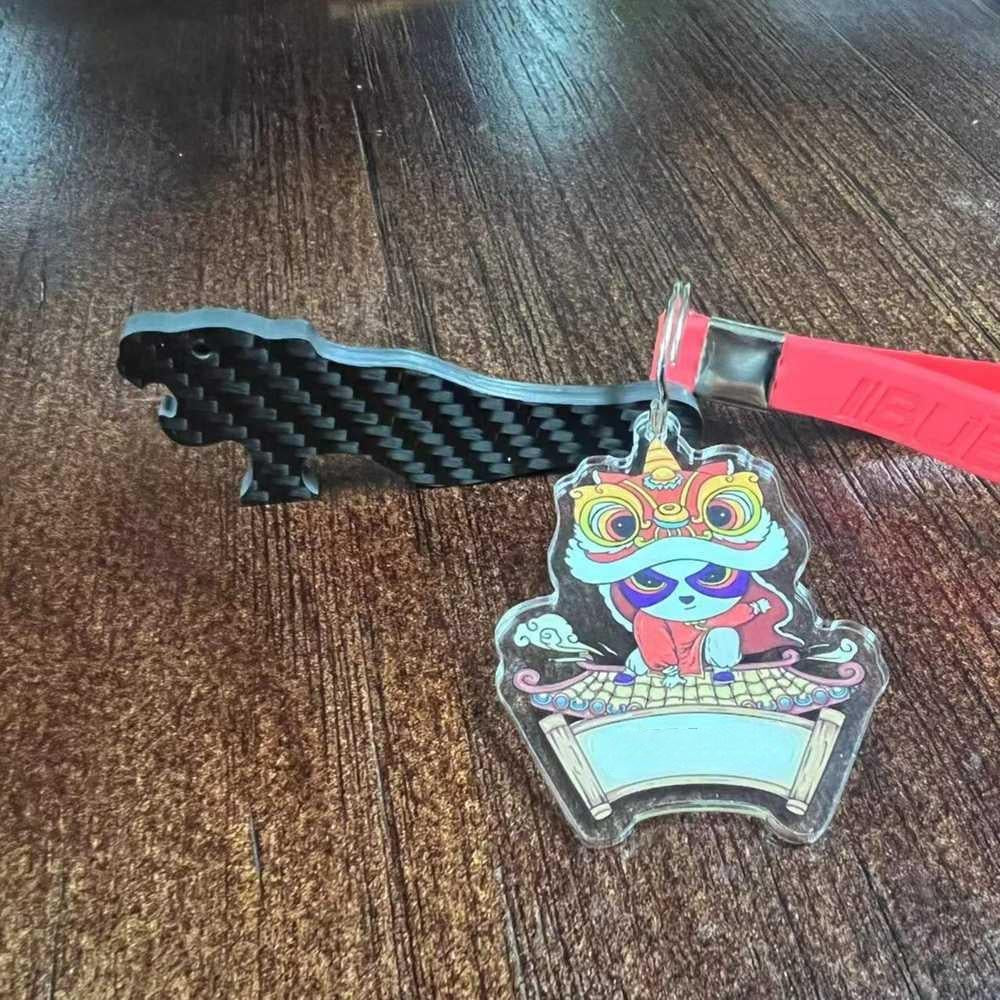 Close-up of a 3K carbon fiber tiger-shaped keychain with a checkered pattern, alongside a colorful acrylic charm featuring a lion dance design with a red strap, placed on a wooden textured surface.