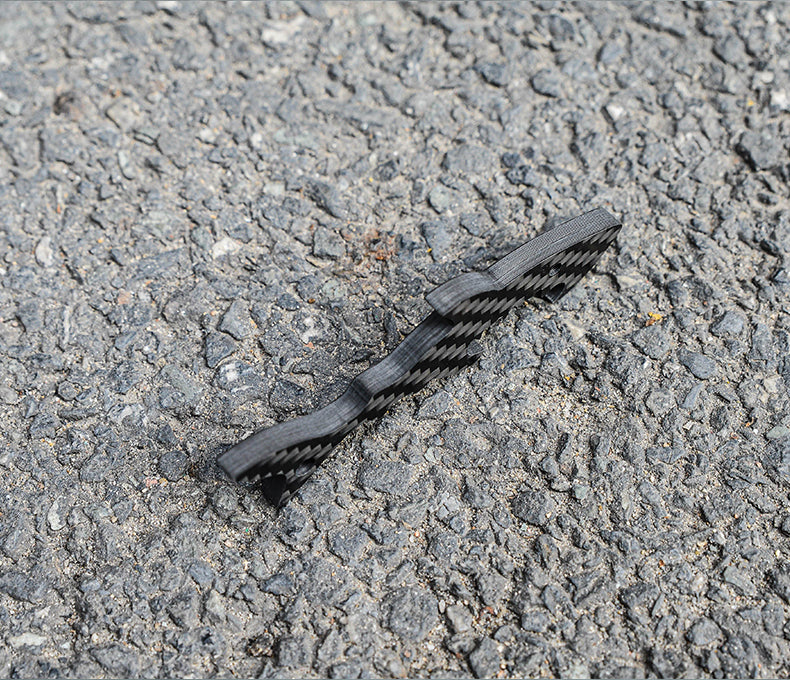 Close-up side view of a shark-shaped 3K carbon fiber keychain with a checkered pattern, placed on a rough asphalt surface.