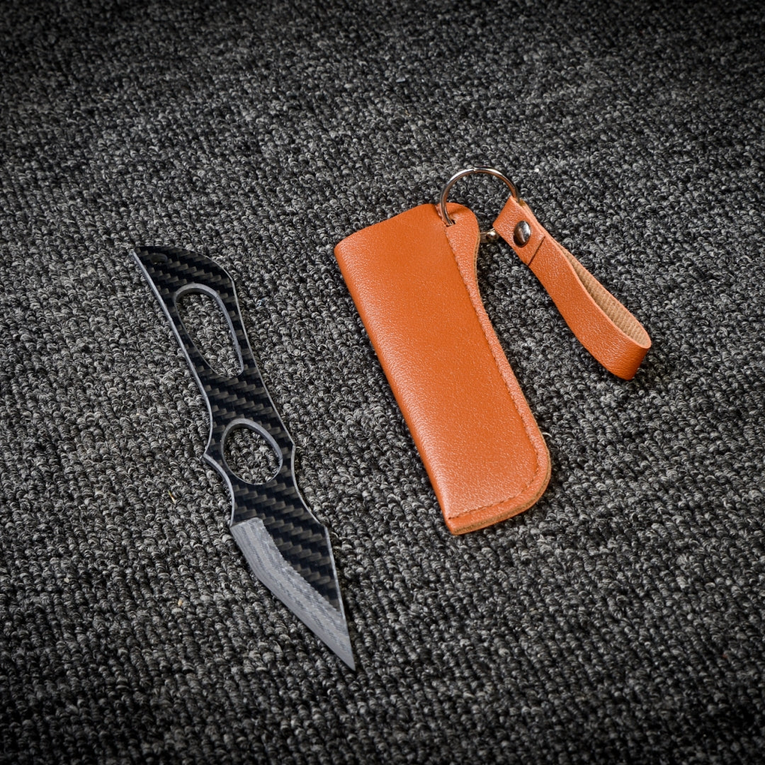 Close-up of a carbon fiber tactical knife with a sleek, patterned design, accompanied by a brown leather sheath with a key ring, displayed on a dark gray textured fabric surface.