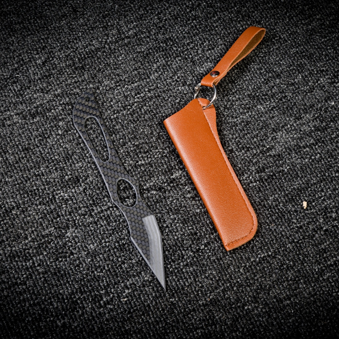 Close-up of a carbon fiber tactical knife with a patterned handle and blade, paired with a brown leather sheath featuring a key ring and strap, displayed on a dark textured fabric surface.