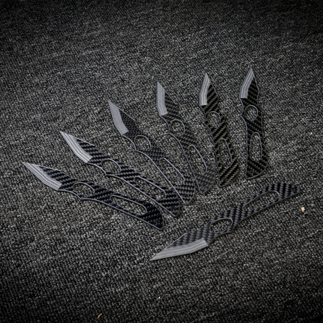 Close-up of multiple carbon fiber throwing knives with distinctive patterned handles and blades, arranged on a dark textured fabric surface.
