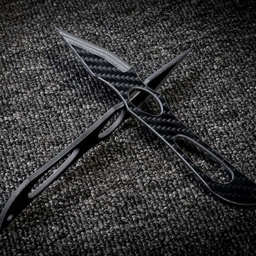 Close-up of two carbon fiber knives with intricate cutout designs and a checkered pattern, crossed over each other on a dark textured fabric surface.
