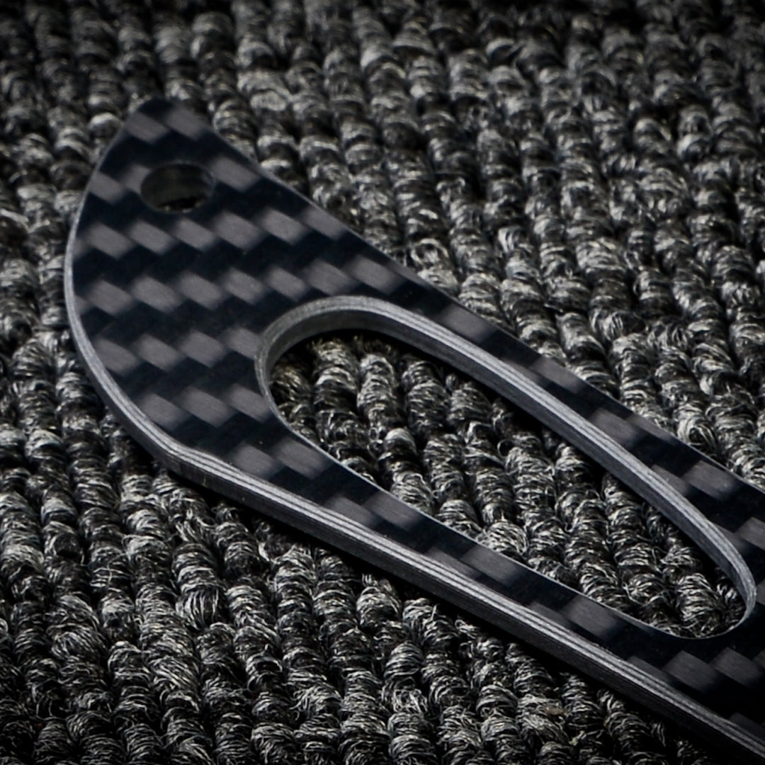 Close-up of a 3K carbon fiber knife handle with a checkered pattern and cutout detail, placed on a dark textured fabric surface.