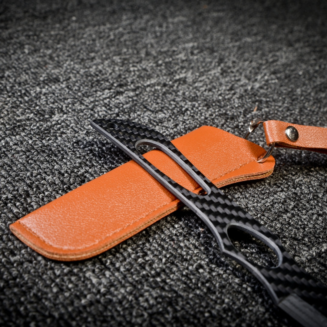 Close-up of a 3K carbon fiber knife with a checkered pattern and cutout details, paired with a brown leather sheath featuring a key ring, displayed on a dark textured fabric surface.