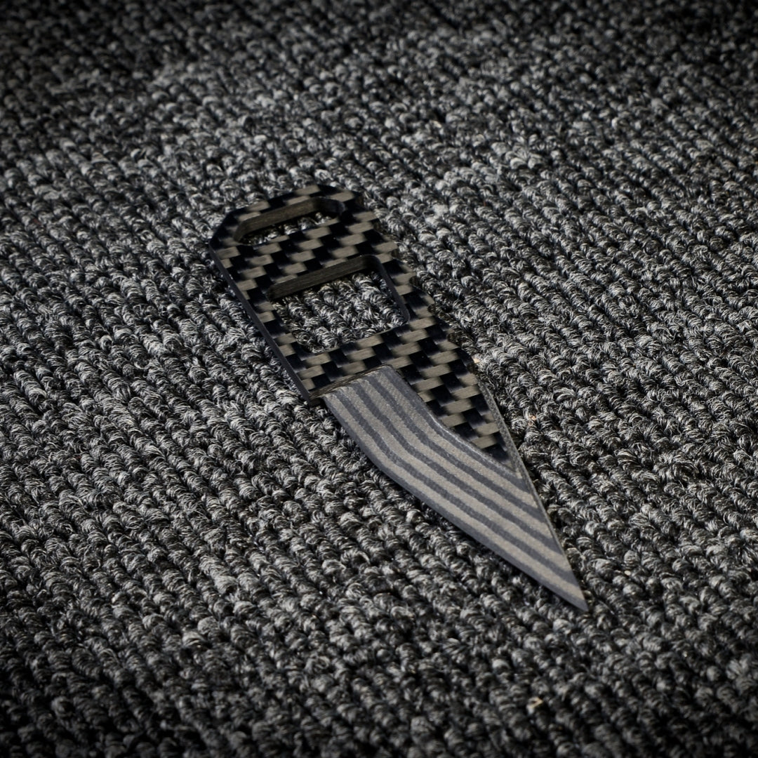 Close-up of a 3K carbon fiber knife with a checkered pattern handle and a striped metallic blade, placed on a dark textured fabric surface.