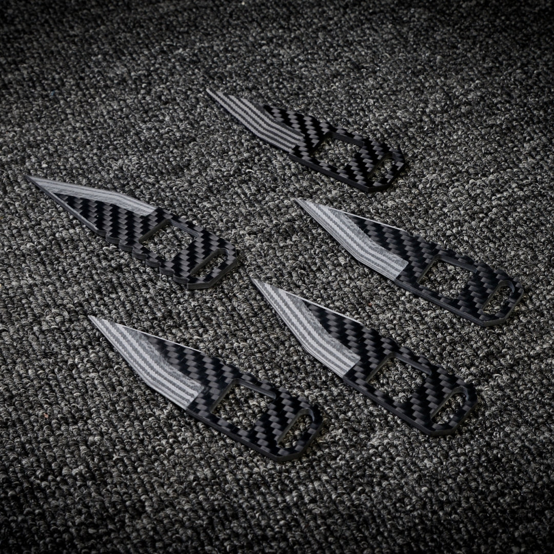 Close-up of five 3K carbon fiber knives with checkered patterns, striped blades, and cutout details, arranged on a dark textured fabric surface.