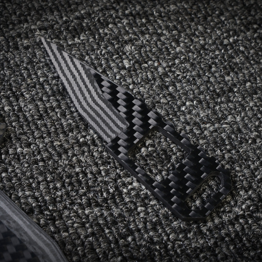 Close-up of a 3K carbon fiber knife featuring a checkered pattern, striped blade, and cutout details on the handle, positioned on a dark textured fabric surface.
