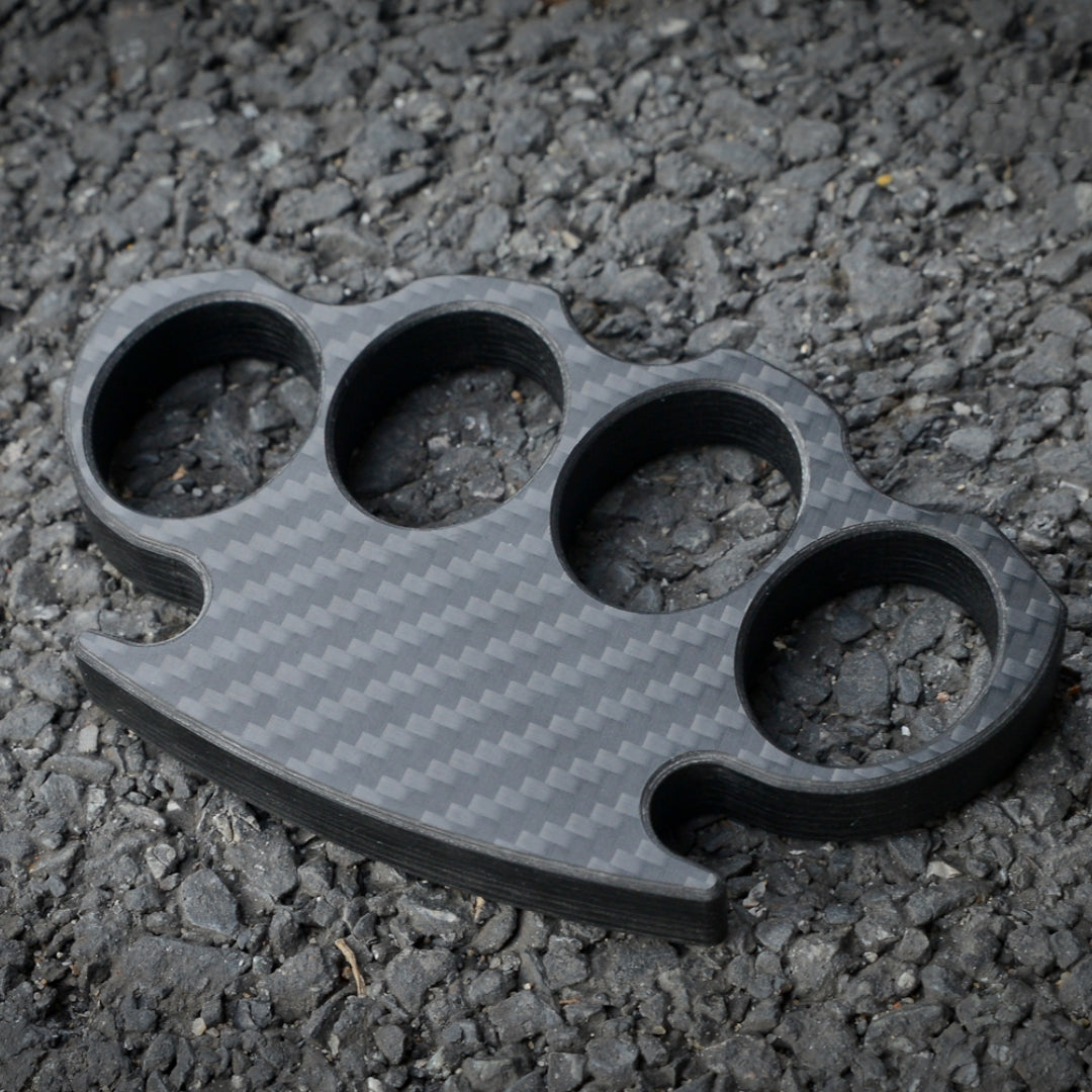 Close-up of a carbon fiber brass knuckles with four finger holes, showcasing its distinct woven pattern, placed on a gravel surface.