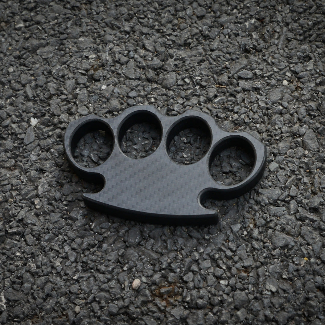 Close-up of a carbon fiber brass knuckles with four finger holes, showcasing its distinct woven pattern, placed on a gravel surface.