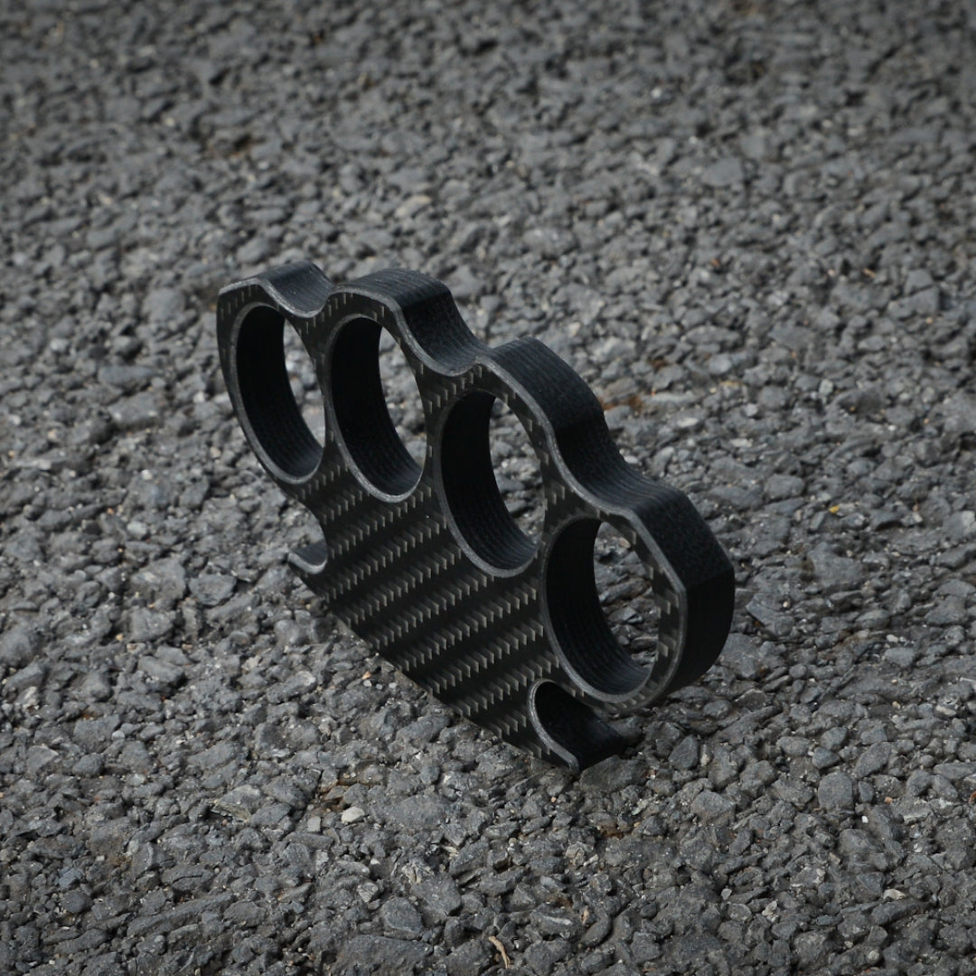 Close-up of a carbon fiber brass knuckles with four finger holes, showcasing its distinct woven pattern, placed on a gravel surface.
