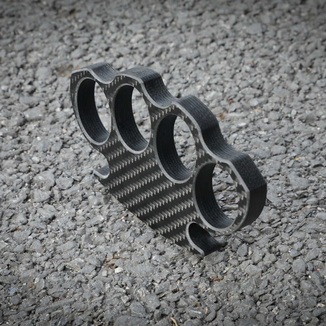 Close-up of a carbon fiber brass knuckles with four finger holes, showcasing its distinct woven pattern, placed on a gravel surface.