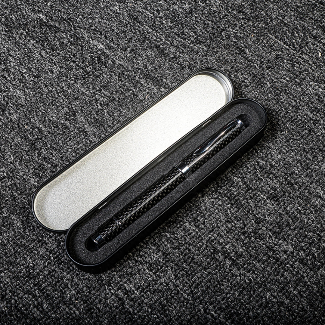 Close-up of a carbon fiber ballpoint pen with a distinct woven texture, nestled in a black metal case with foam lining and a silver interior, displayed on a gray textured fabric surface.