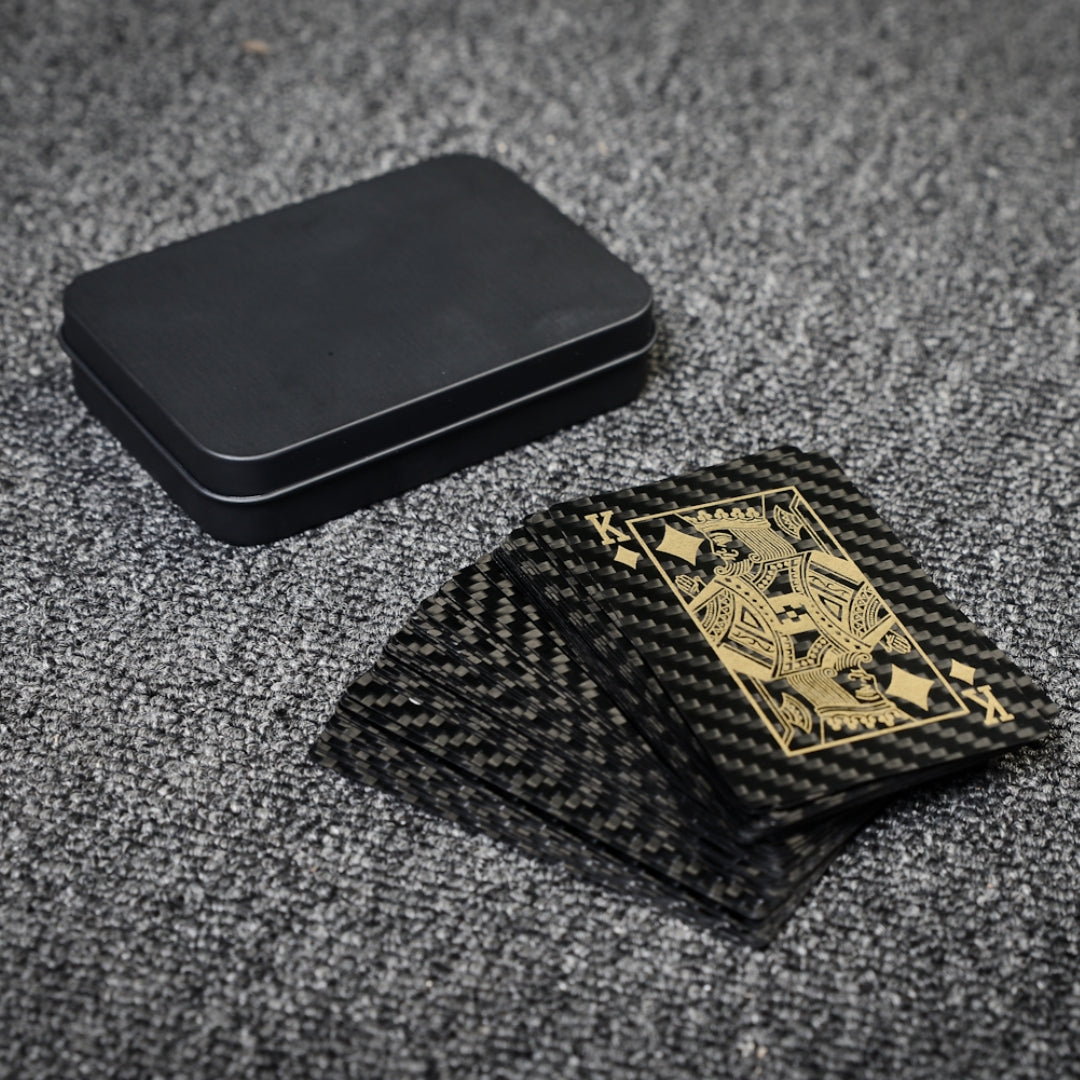 Close-up of carbon fiber playing cards with gold detailing (featuring the King of Diamonds) and a black tin case, laid on a gray textured surface.