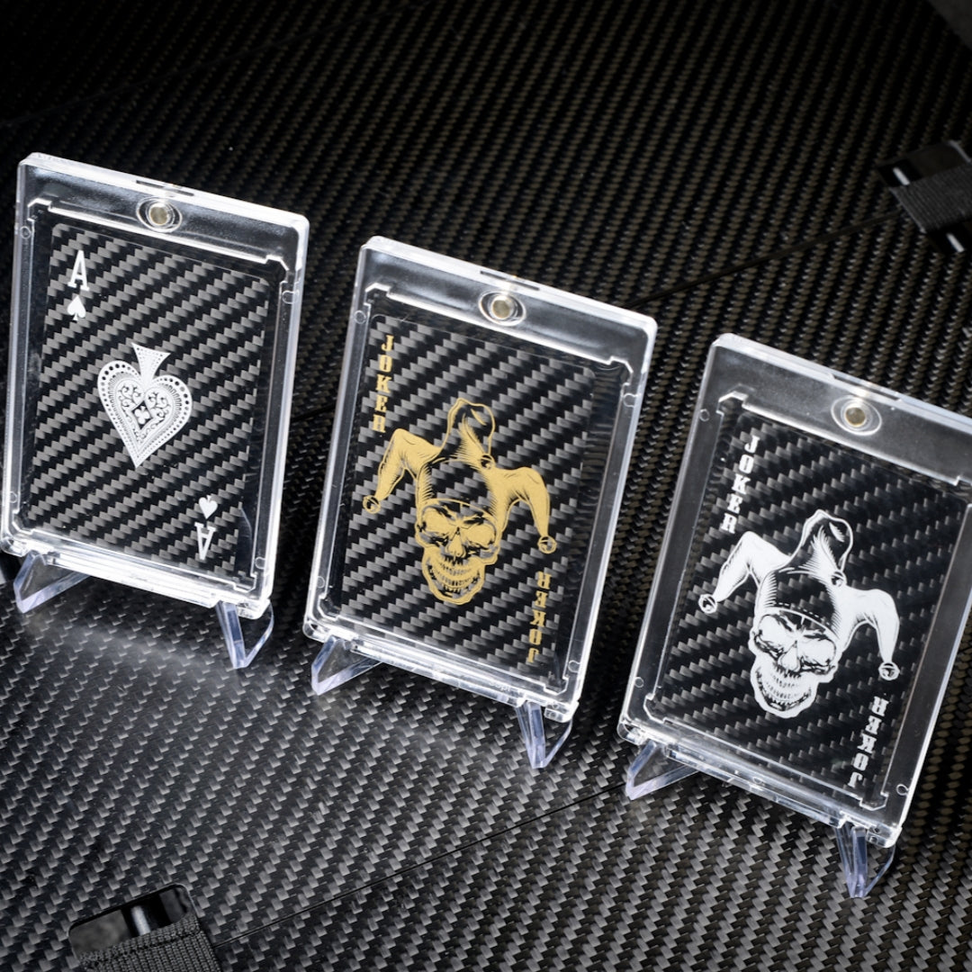 Close-up of three carbon fiber playing cards (Ace of Spades, gold Joker, and white Joker) in clear protective cases, displayed on a carbon fiber textured surface.