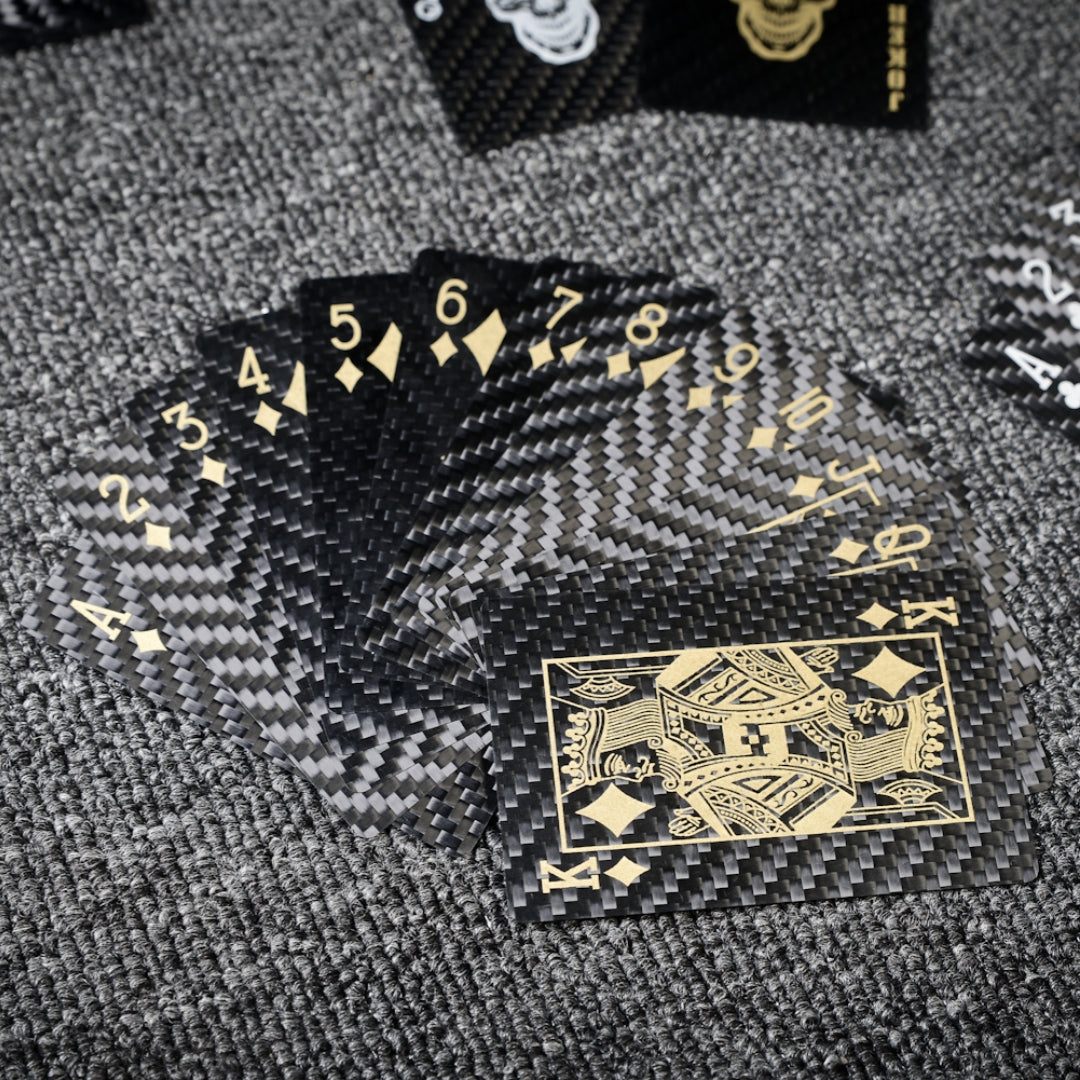 Close-up of carbon fiber playing cards with gold diamond suit details (including the King of Diamonds), fanned out on a gray textured surface.