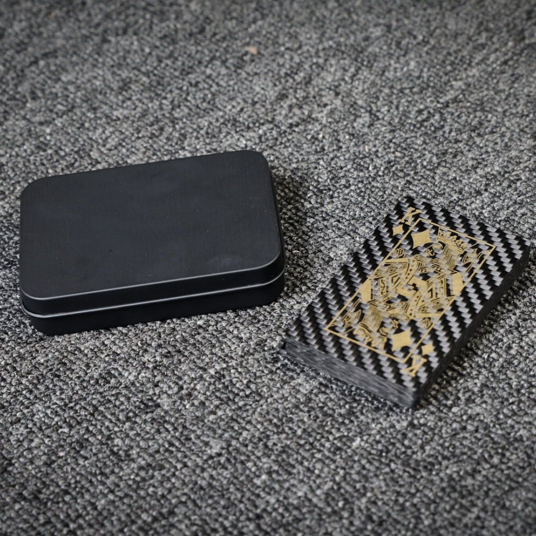 Close-up of a deck of carbon fiber playing cards with gold detailing (featuring the King card) and a black tin case, laid on a gray textured surface.
