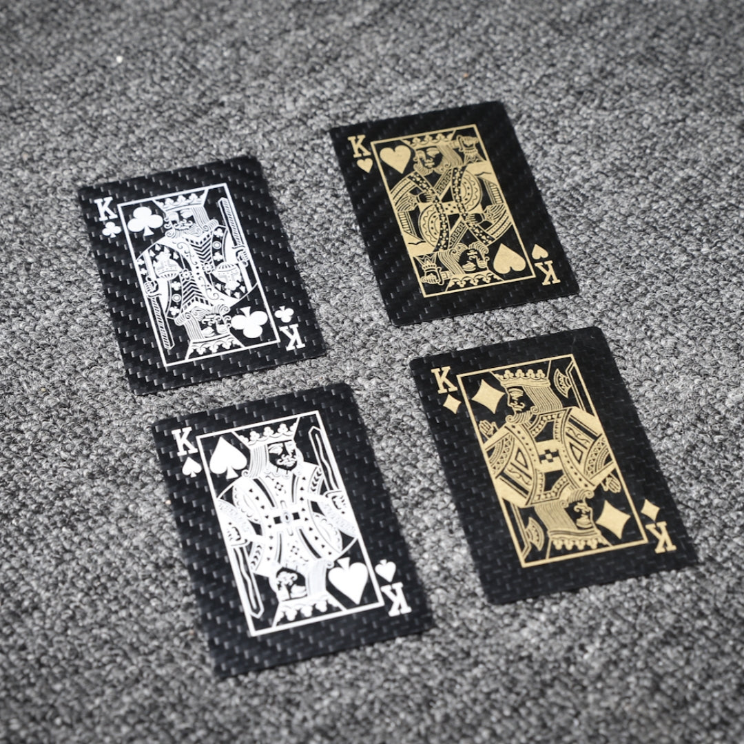 Close-up of four carbon fiber playing cards (Kings of Clubs, Hearts, Spades, and Diamonds) featuring white and gold detailing, laid on a gray textured surface.