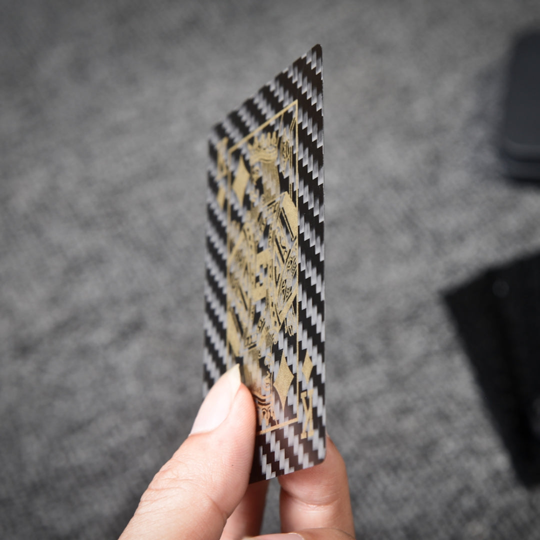 Close-up of a carbon fiber playing card (King of Diamonds) with gold detailing, held by a hand, showcasing the distinct carbon fiber weave pattern against a gray textured background.