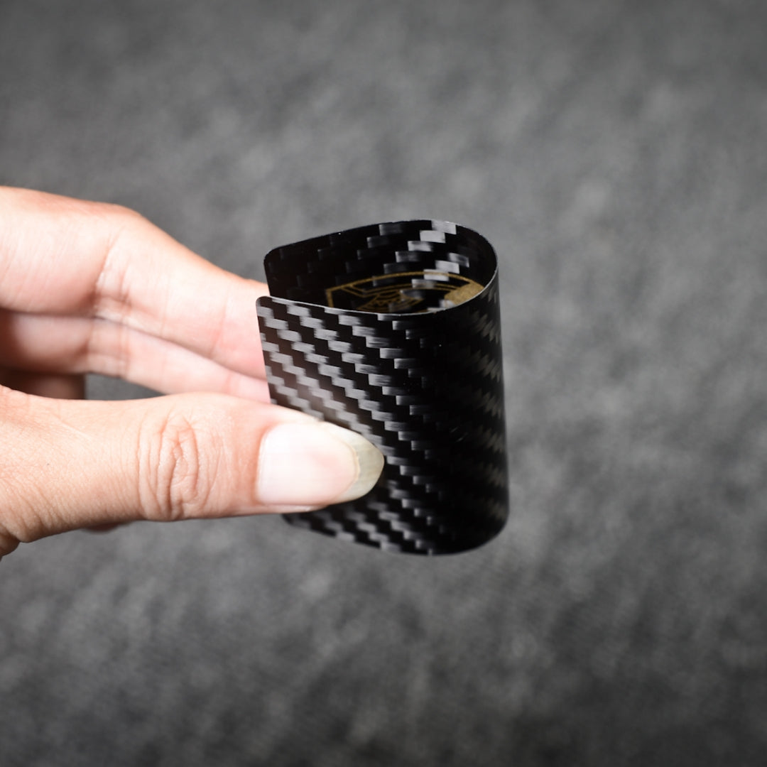 Close-up of a carbon fiber playing card with gold detailing, bent to showcase its flexibility, held by a hand against a gray background, highlighting the distinct carbon fiber weave pattern.