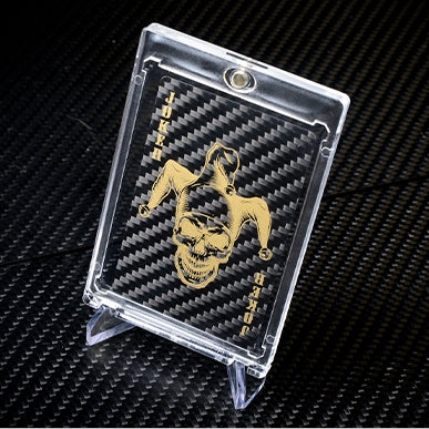 Close-up of a carbon fiber playing card with a gold Joker skull-jester design, encased in a clear protective case and displayed on a carbon fiber textured surface.