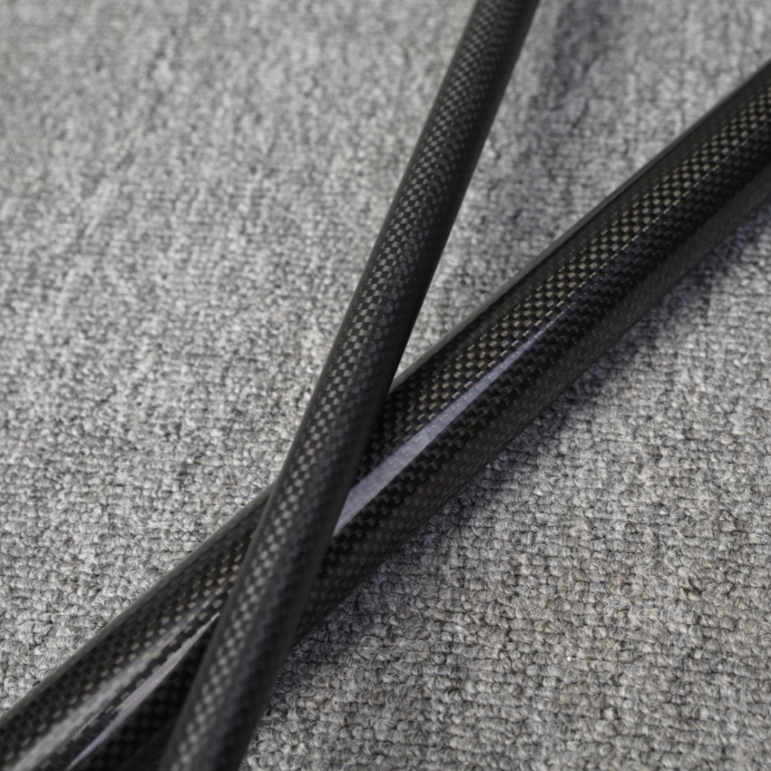 Close-up detail of two crossed sections of a carbon fiber pool cue, showcasing the textured carbon fiber pattern, placed on a gray fabric surface.