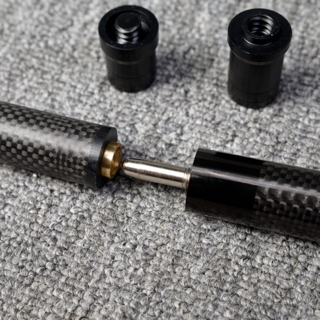Close-up detail of a two-piece carbon fiber pool cue with a textured carbon fiber pattern, showcasing the metal joint and brass fitting connection, along with two black accessories, placed on a gray textured surface.
