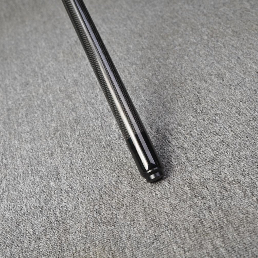 Close-up detail of the end of a carbon fiber pool cue, showcasing a textured carbon fiber pattern and a smooth black finish at the tip, placed on a gray textured surface.