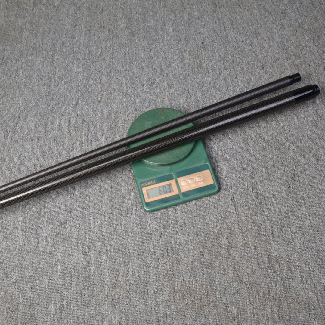 Close-up of two carbon fiber pool cues placed on a digital scale displaying a weight reading, set against a gray textured surface.
