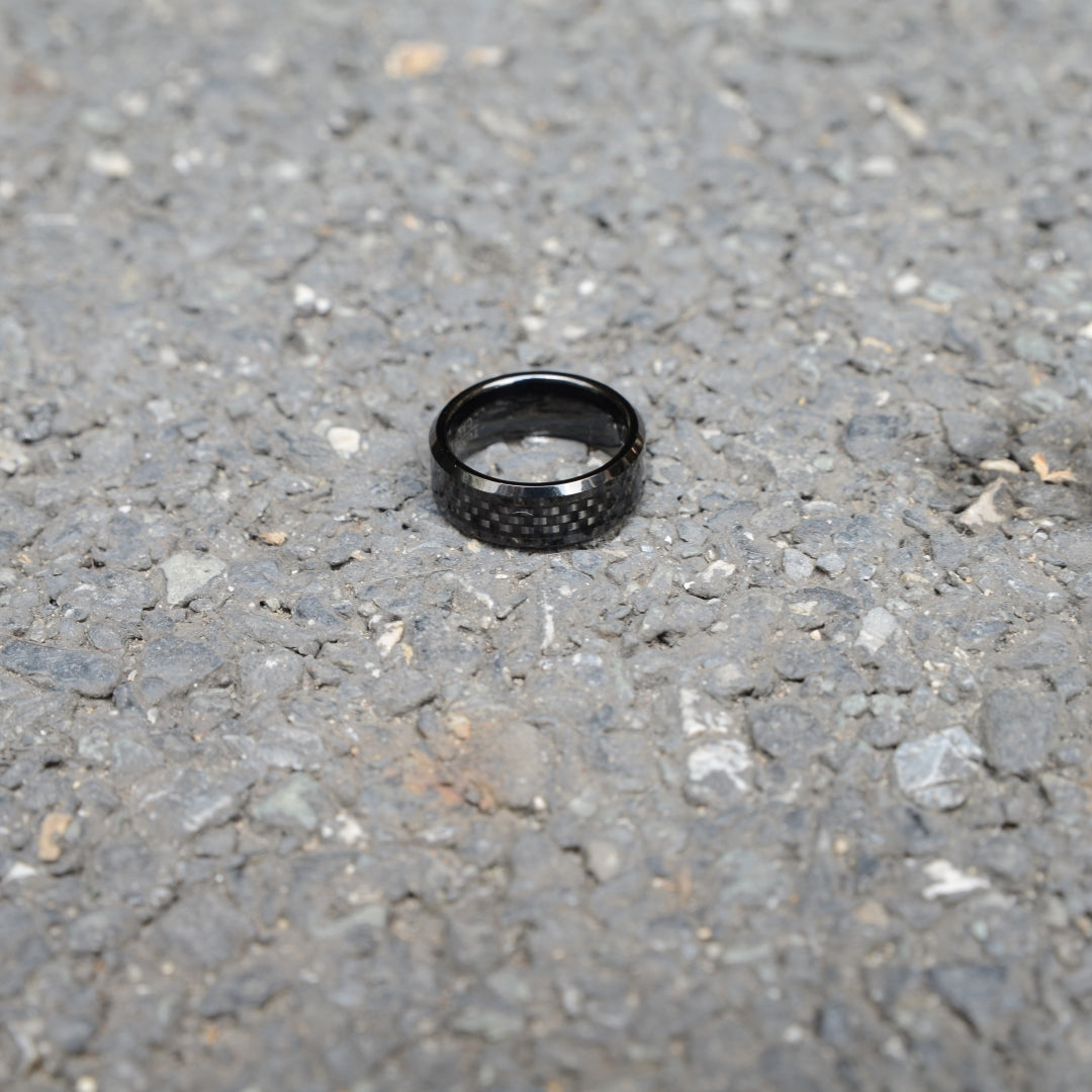 A black ring featuring 3K carbon fiber detailing, resting on a rough gravel surface.